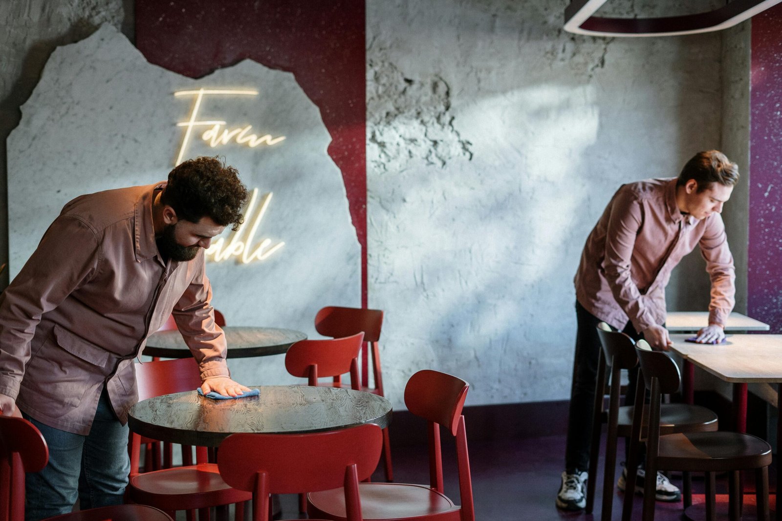 Two staff members cleaning tables in a modern café with neon signage and red chairs.